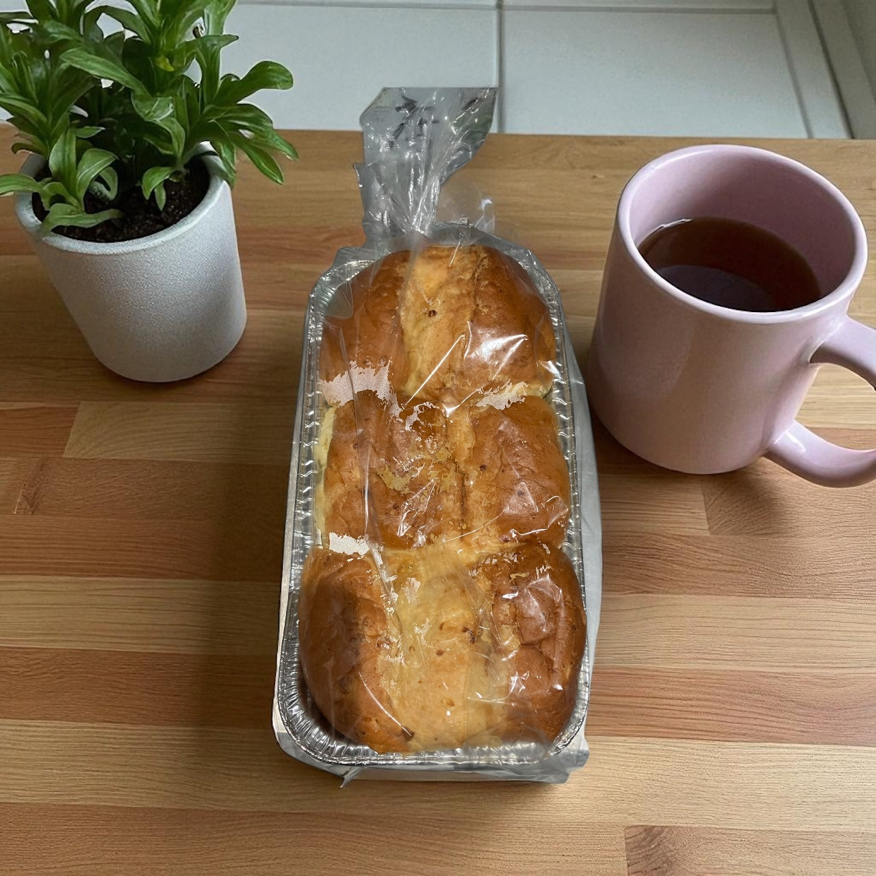 Wrapped corn bread loaf in aluminum tray on wooden table with pink mug and small potted plant nearby